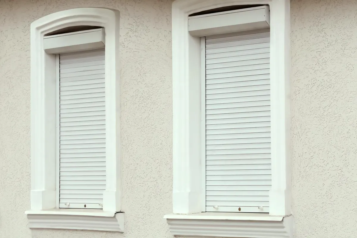 Deux volets roulants blancs fermés, coffres posés dans l'embrasure de fenêtres à encadrements moulurés sur une façade claire